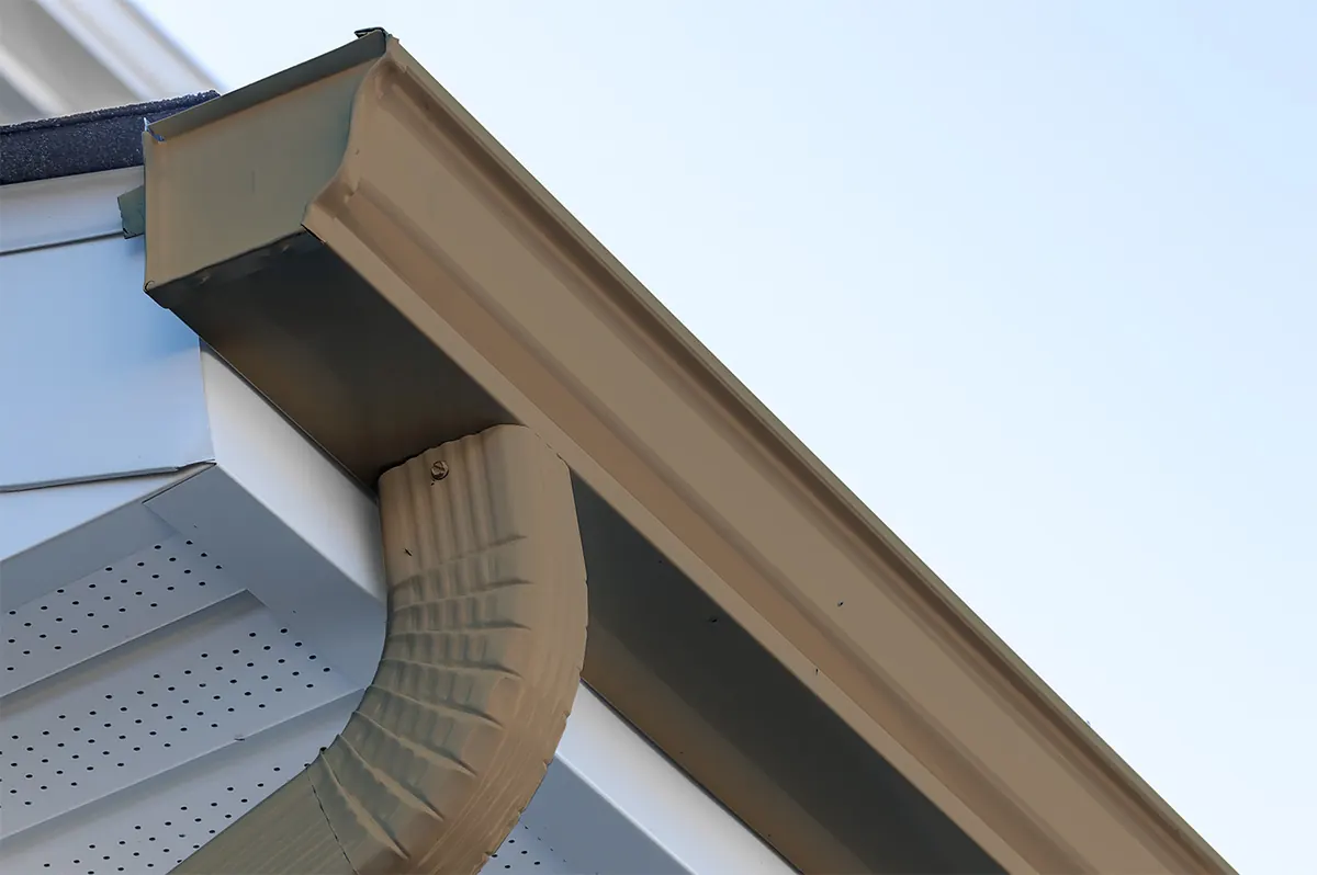 Beige aluminum gutter and curved downspout at a roof edge